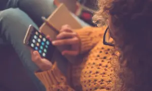 Kobieta w okularach i swetrze trzyma smartfon z ikonami aplikacji i notatnik.