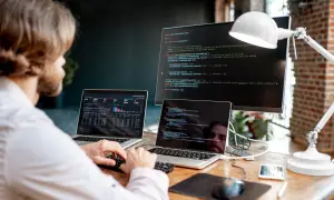 Programista pracuje przy biurku z dwoma laptopami i monitorem, na których widać kod i wykresy.