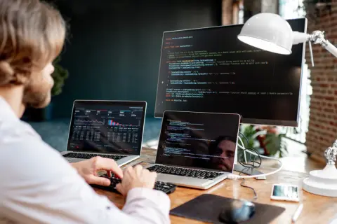 Programista pracuje przy biurku z dwoma laptopami i monitorem, na których widać kod i wykresy.