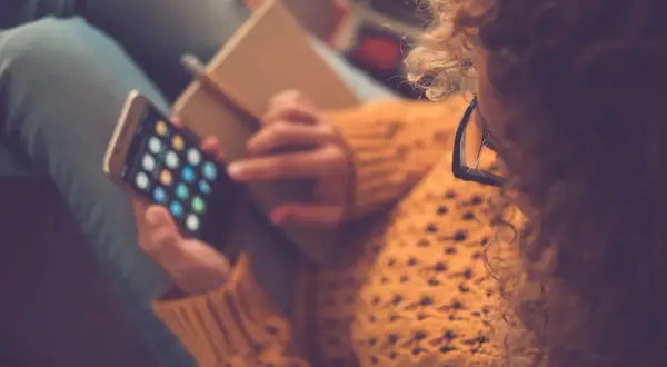 Kobieta w okularach i swetrze trzyma smartfon z ikonami aplikacji i notatnik.