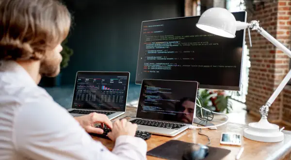 Programista pracuje przy biurku z dwoma laptopami i monitorem, na których widać kod i wykresy.