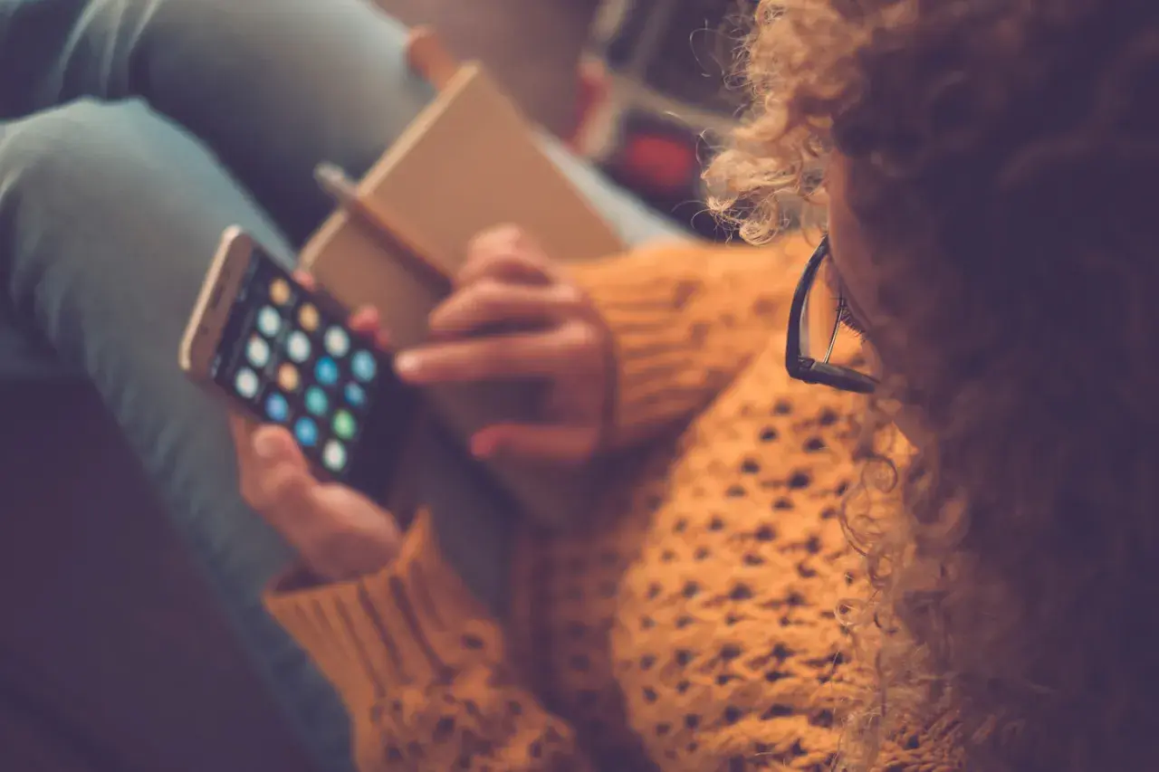 Kobieta w okularach i swetrze trzyma smartfon z ikonami aplikacji i notatnik.