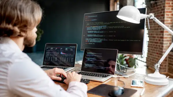 Programista pracuje przy biurku z dwoma laptopami i monitorem, na których widać kod i wykresy.